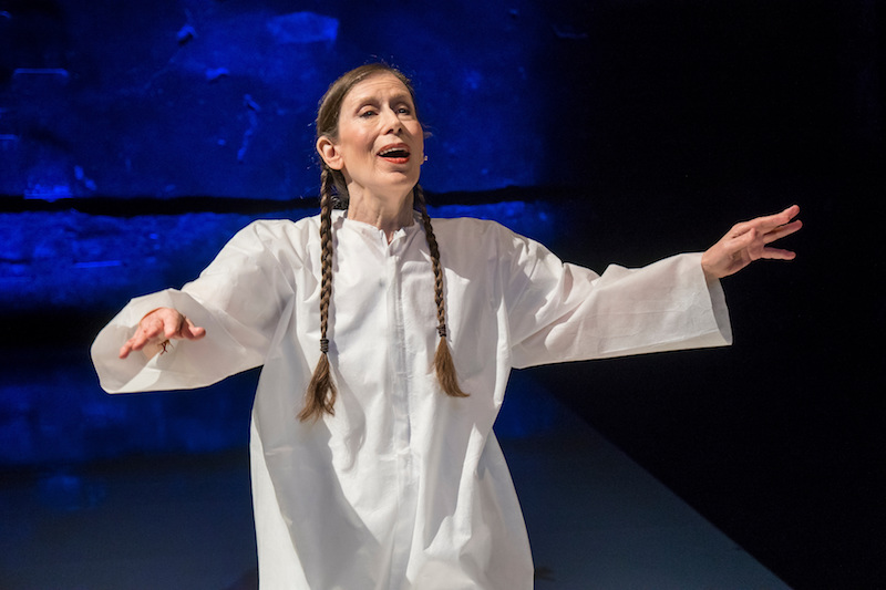 Meredith Monk, in a white caftan and her hair in two braids, sings with her arms out to her sides.